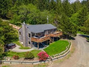 A modern two-story house with a deck, surrounded by lush greenery and a dirt road. - Headland- Oceanfront Family Friendly (Gold Beach)