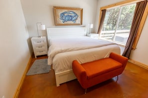 A bedroom with a large bed, white nightstands, a red bench, a painting of a crab, and a window with curtains. - Headland- Oceanfront Family Friendly (Gold Beach)