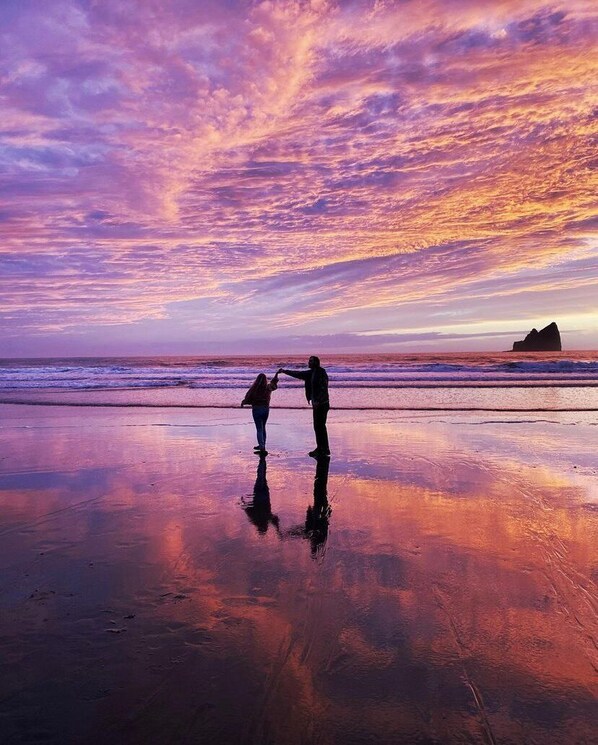 A beach scene with two people holding hands, a rocky outcrop in the distance, and a sunset sky.
