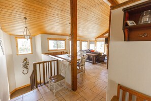 A spacious kitchen with wooden beams, a dining area with white chairs, and a view of the outdoors through large windows.