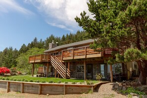 A two-story wooden cabin with a wooden deck and staircase, surrounded by a landscaped garden and trees.