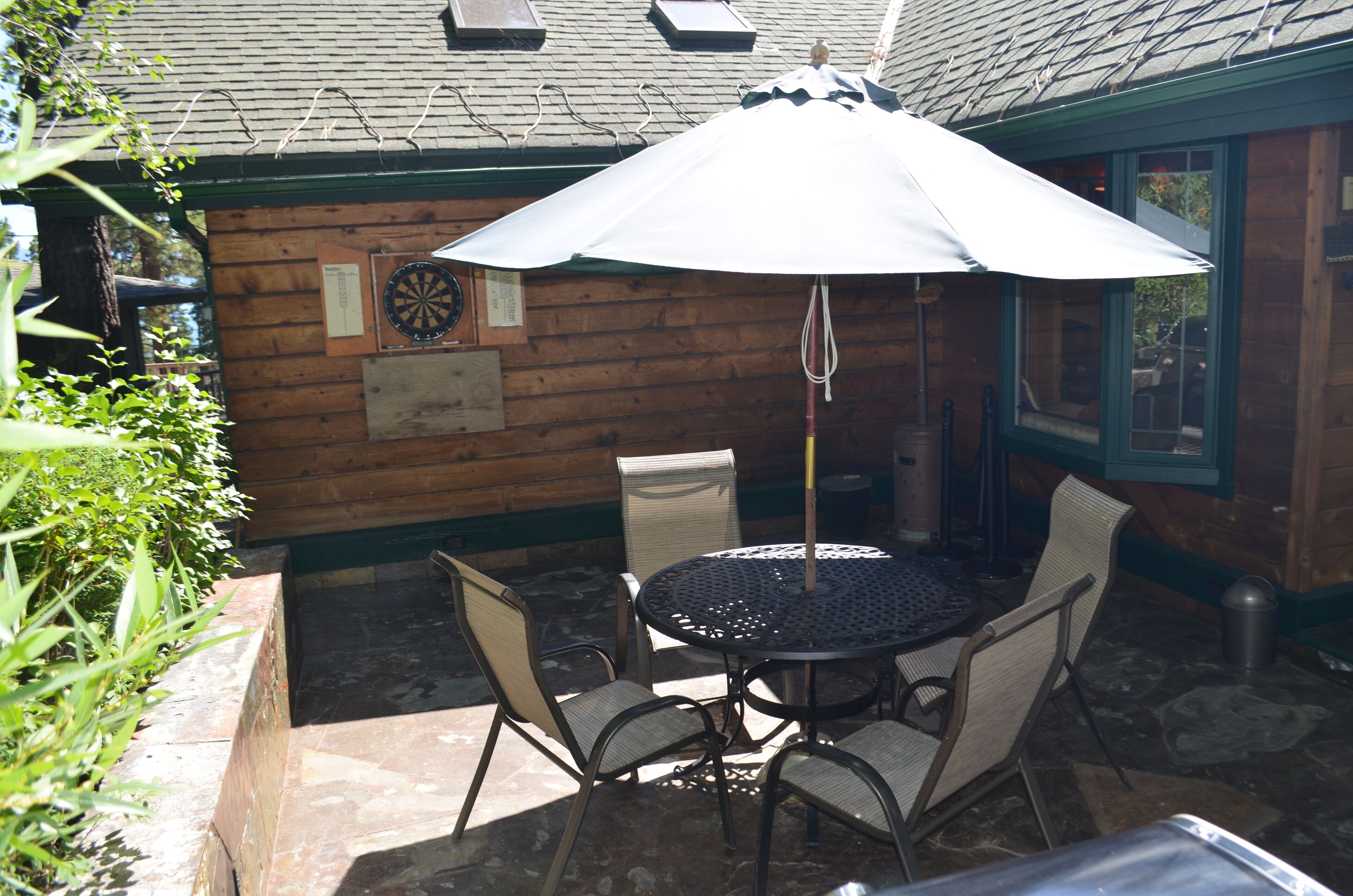 Eine Terrasse mit einem runden Tisch, vier Stühlen, einem Sonnenschirm und einer Dartscheibe an einer Holzwand.