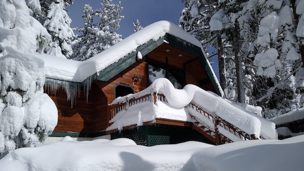 Una cabaña de madera con techo inclinado cubierto de nieve, rodeada de árboles cargados de nieve.
