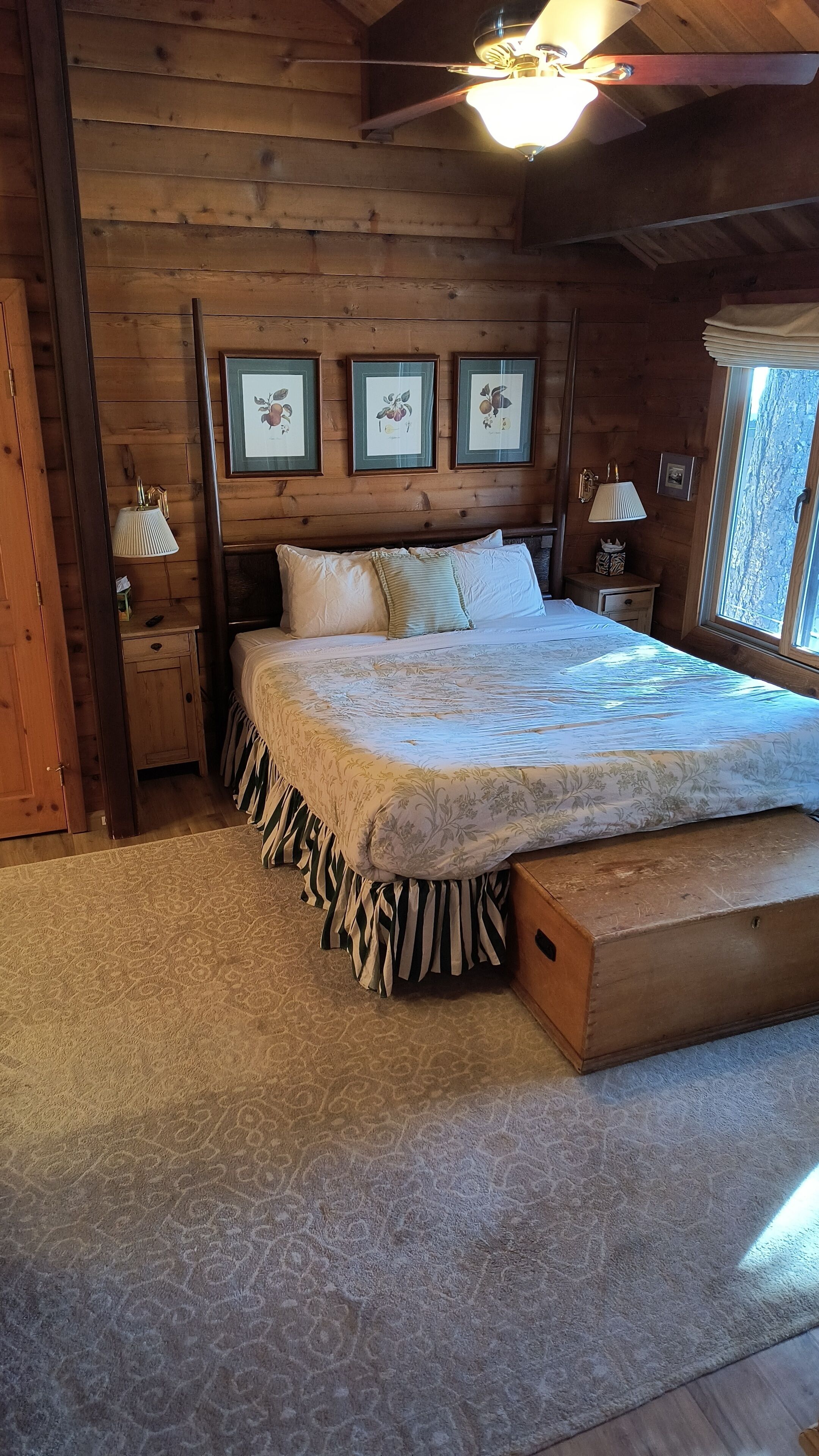 Une chambre confortable dans un chalet en bois, avec un grand lit, des reproductions botaniques encadrées accrochées au mur et une fenêtre offrant une belle vue.