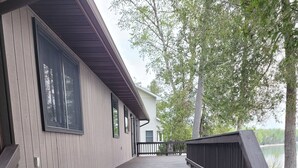 Eine Holzterrasse führt zu einem Haus mit schwarzer Geländer, umgeben von Bäumen und einem Gewässer.