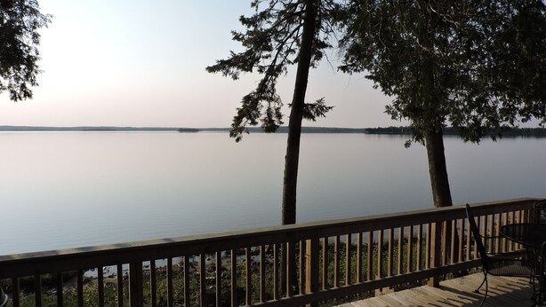 Blick auf einen See von einer Holzterrasse mit Geländer, mit ruhigem Wasser und Bäumen in der Ferne.