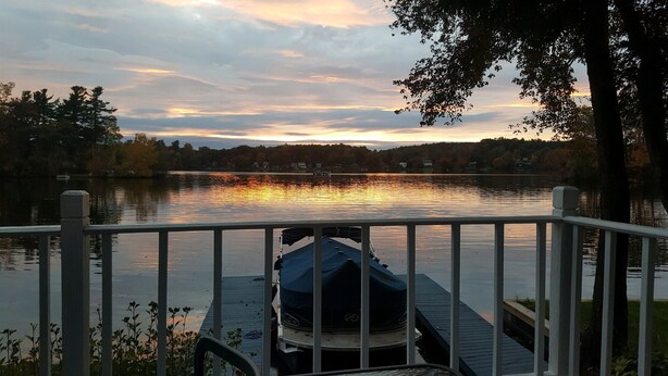 A dock with a covered boat, a wooden railing, and a view of a lake with trees and a sunset in the background. - Heaven Can't Wait, On Beautiful Kinderhook Lake!! (Valatie)
