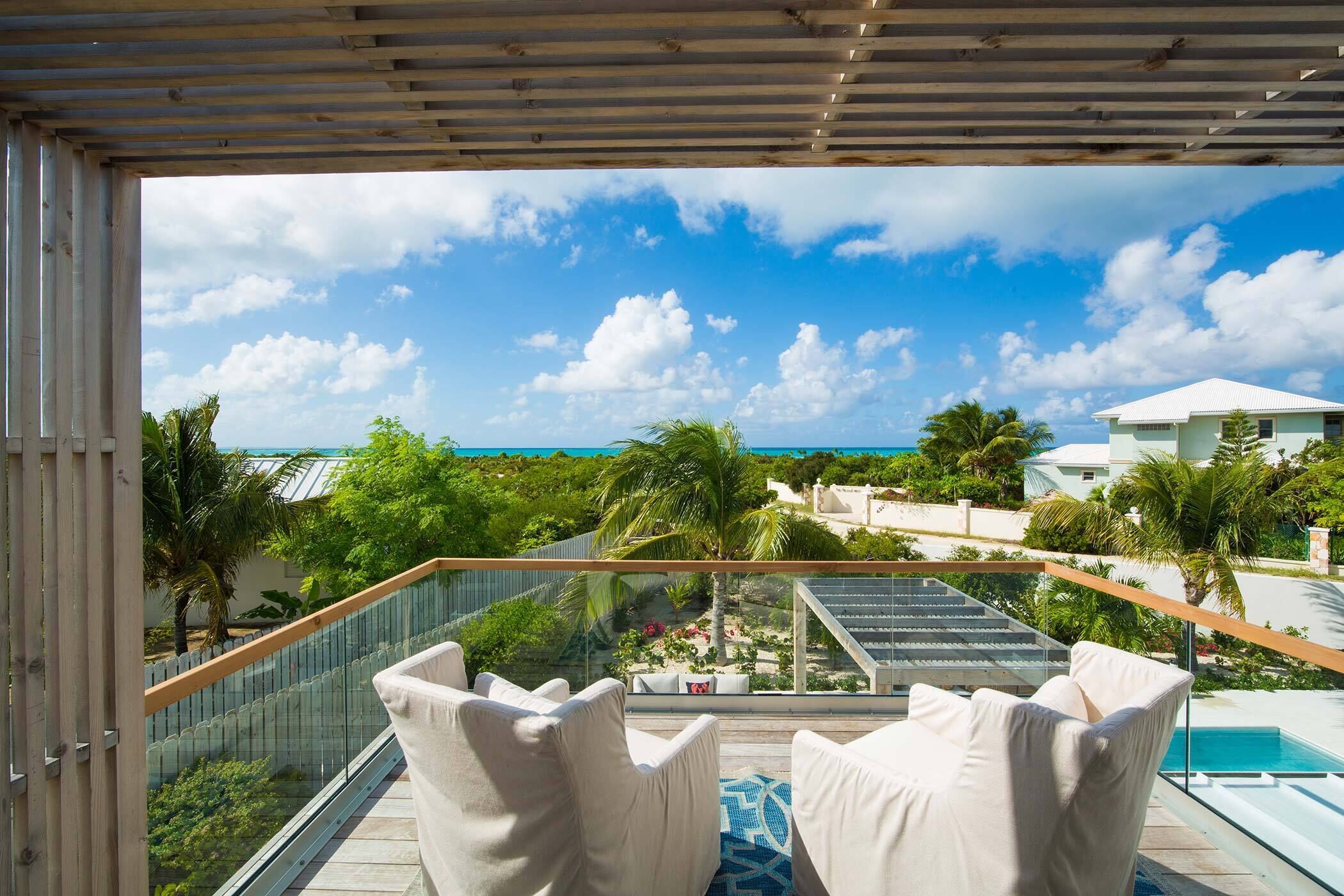 A balcony with white cushioned chairs, a glass railing, and a view of the ocean and palm trees.