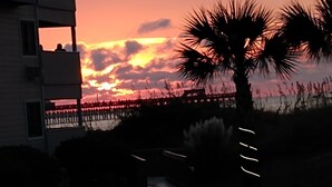 Una vista del atardecer con un muelle que se adentra en el agua, palmeras y un edificio con balcones.
