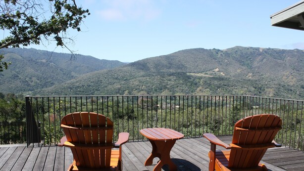 Um deck de madeira com duas cadeiras Adirondack e uma mesa pequena, com vista para uma paisagem montanhosa.