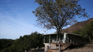 Uma casa térrea com varanda, rodeada por árvores e colinas.