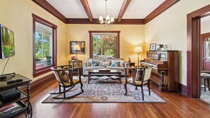 A spacious living room with a wooden floor, a large window, a piano, and a chandelier. - The St. Helena House (Saint Helena)