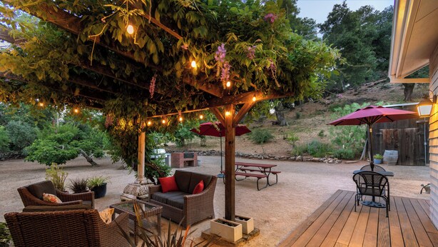 An outdoor patio with wicker furniture, a pergola with string lights, and a red umbrella. - The St. Helena House (Saint Helena)