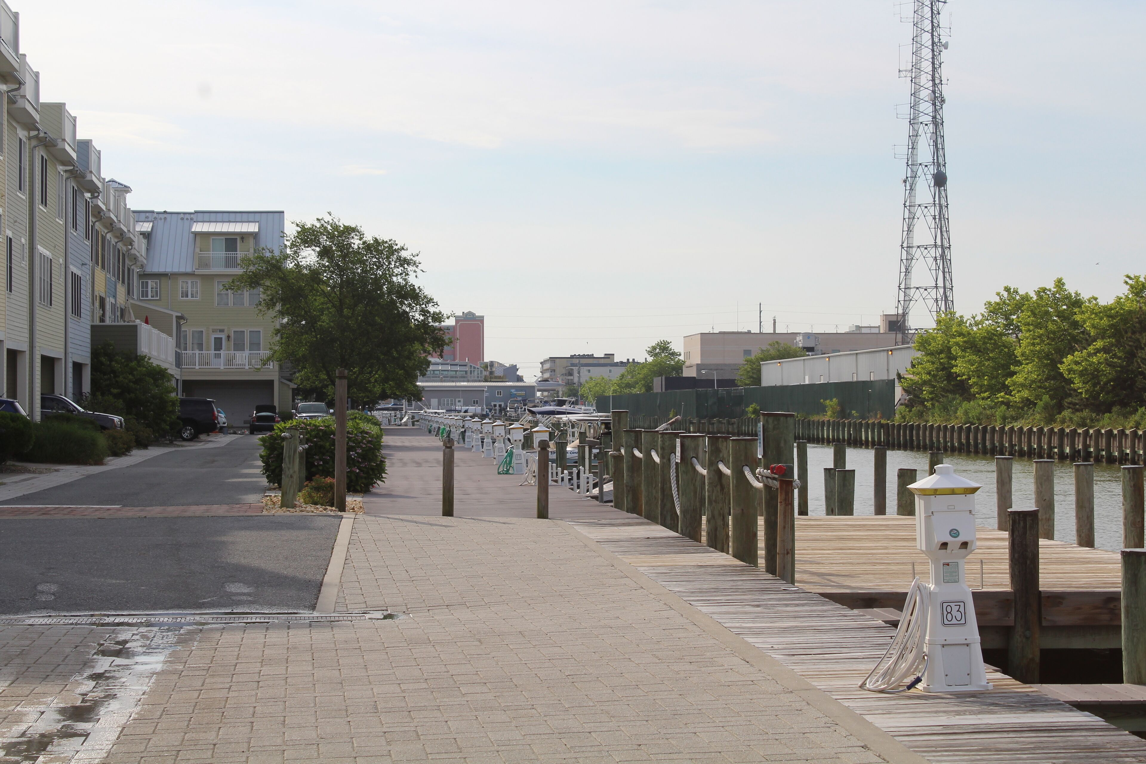 A waterfront promenade with a wooden boardwalk, a row of buildings on one side, and a tall communication tower in the background.