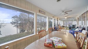 A dining area with a large table, chairs, and a view of a lake. There are books on the table and a ceiling fan above.