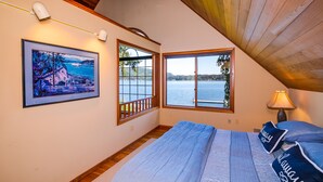 A bedroom with a large bed, two windows with views of a lake, a framed picture on the wall, and a lamp on the nightstand. - Lake Whatcom Beach Resort (Bellingham)