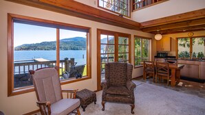 A spacious living room with large windows overlooking a lake, a wooden rocking chair, a patterned armchair, and a dining area with a kitchen in the background. - Lake Whatcom Beach Resort (Bellingham)