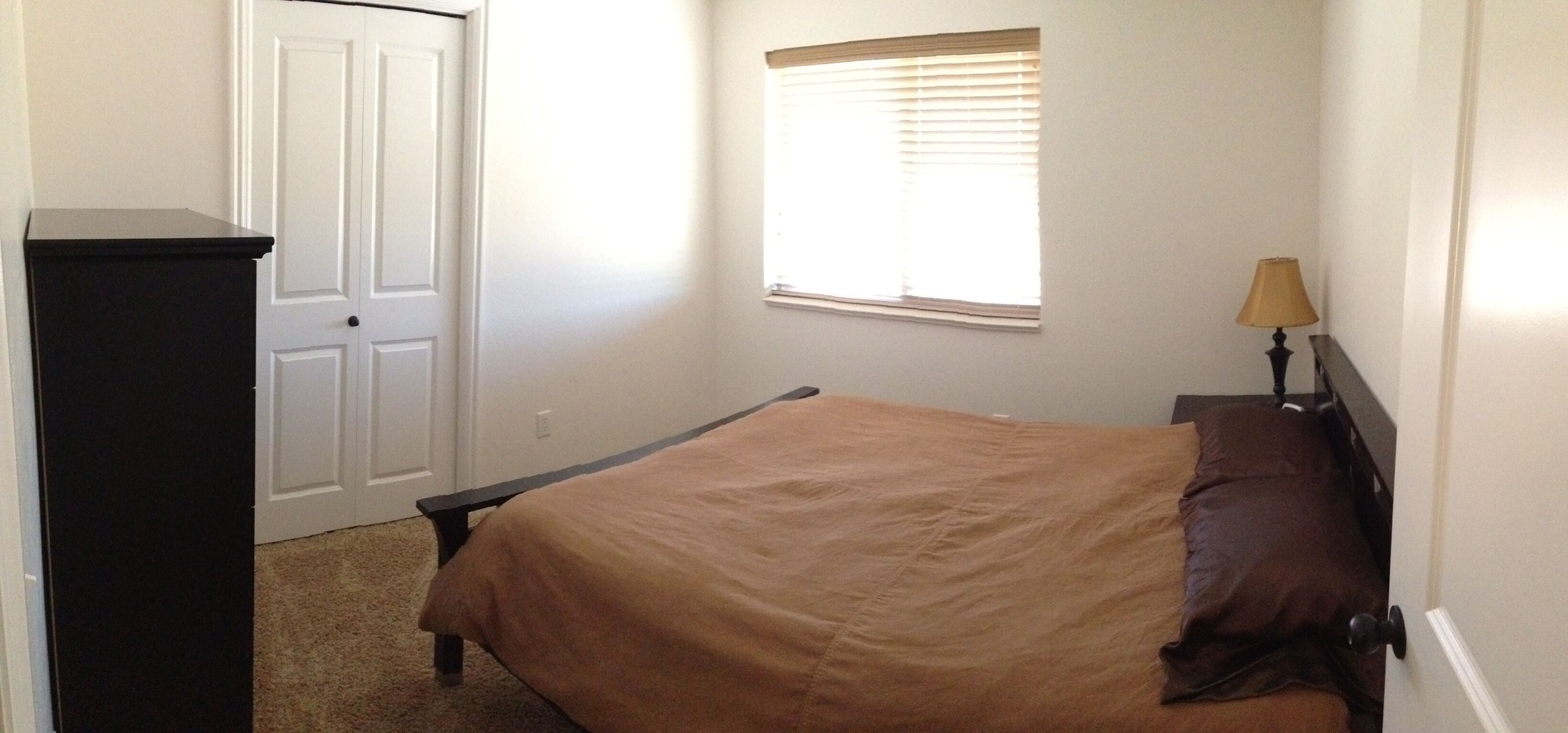 A bedroom with a bed, a dresser, a window with blinds, and a nightstand with a lamp.