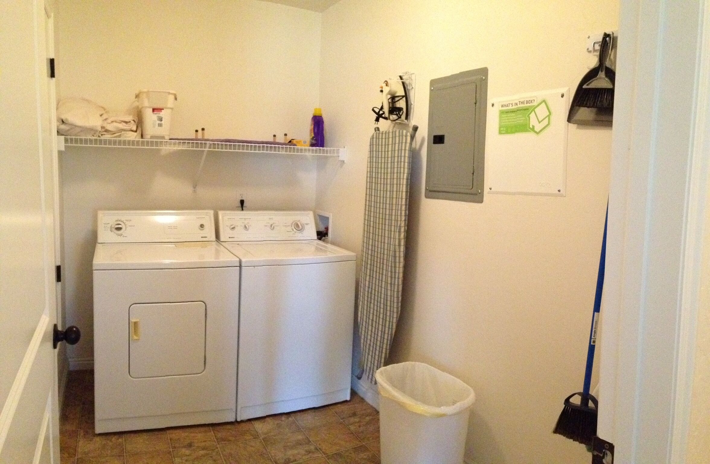 A laundry area with two washing machines, a shelf with cleaning supplies, a broom, and a trash bin.