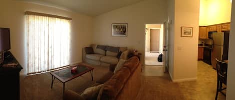 A living room with a sofa, a coffee table, and a television. There is a kitchen area visible through an open doorway.