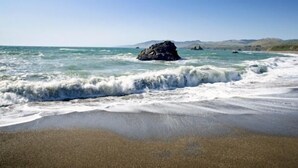 A beach with waves crashing onto the shore, a large rock formation in the water, and distant hills.