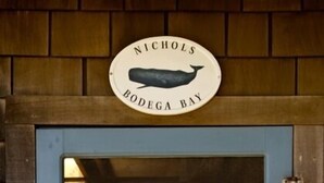 A wooden door with a sign reading "Nichols Bodega Bay" and a view of the ocean through a glass window.