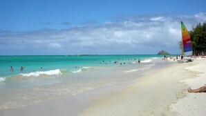 Ein Strand mit klarem türkisblauem Wasser, Menschen beim Schwimmen und Sonnenbaden, und ein buntes Segelboot auf der rechten Seite.