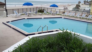 Piscina com espreguiçadeiras e guarda-sóis, cercada por uma grade, com vista para a praia e oceano ao fundo.