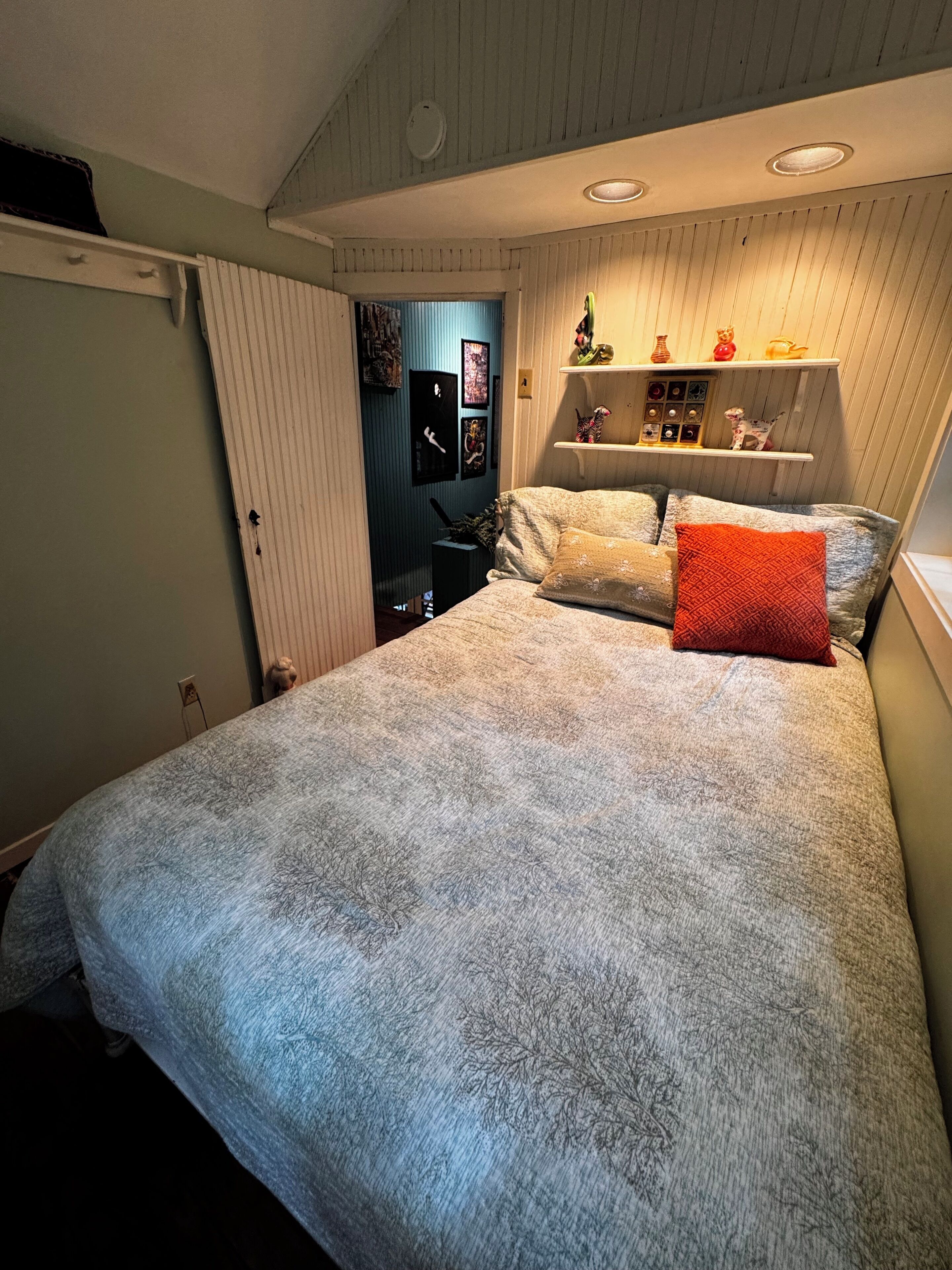 Una camera da letto con un letto, cuscini, una mensola con oggetti decorativi e una porta che dà su un'altra stanza.