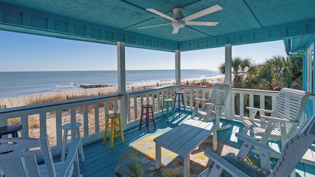 A covered porch with a view of the beach, featuring a ceiling fan, white chairs, a table, and a colorful rug. - 4BR/4Bth Direct Oceanfront,House,Covered Porch and fireplace! (Edisto Beach)