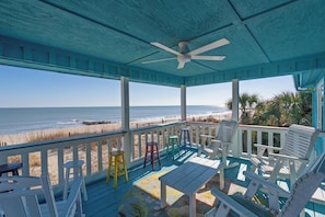 A covered porch with a view of the beach, featuring a ceiling fan, white chairs, a table, and a colorful rug.