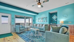 A living room with teal walls, a ceiling fan, a patterned rug, and a view of the ocean through large windows. - 4BR/4Bth Direct Oceanfront,House,Covered Porch and fireplace! (Edisto Beach)