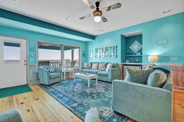 A living room with teal walls, a ceiling fan, a patterned rug, and a view of the ocean through large windows.