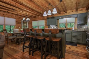 A rustic cabin interior with wooden floors, a dining area with stools, a kitchen with green cabinets, and a large window overlooking greenery.