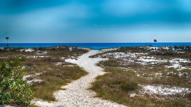 Ein sandiger Pfad führt zu einem Strand mit Dünen und blauem Himmel.