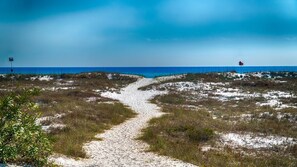 Un camí de sorra que porta a una platja amb dunes i un cel blau clar.