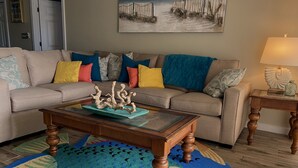 Ein Wohnzimmer mit einem beige farbenen Sofa, bunten Kissen, einem Couchtisch aus Holz mit einem dekorativen Gegenstand, einem Teppich in Blau- und Grüntönen und einem Bild an der Wand.