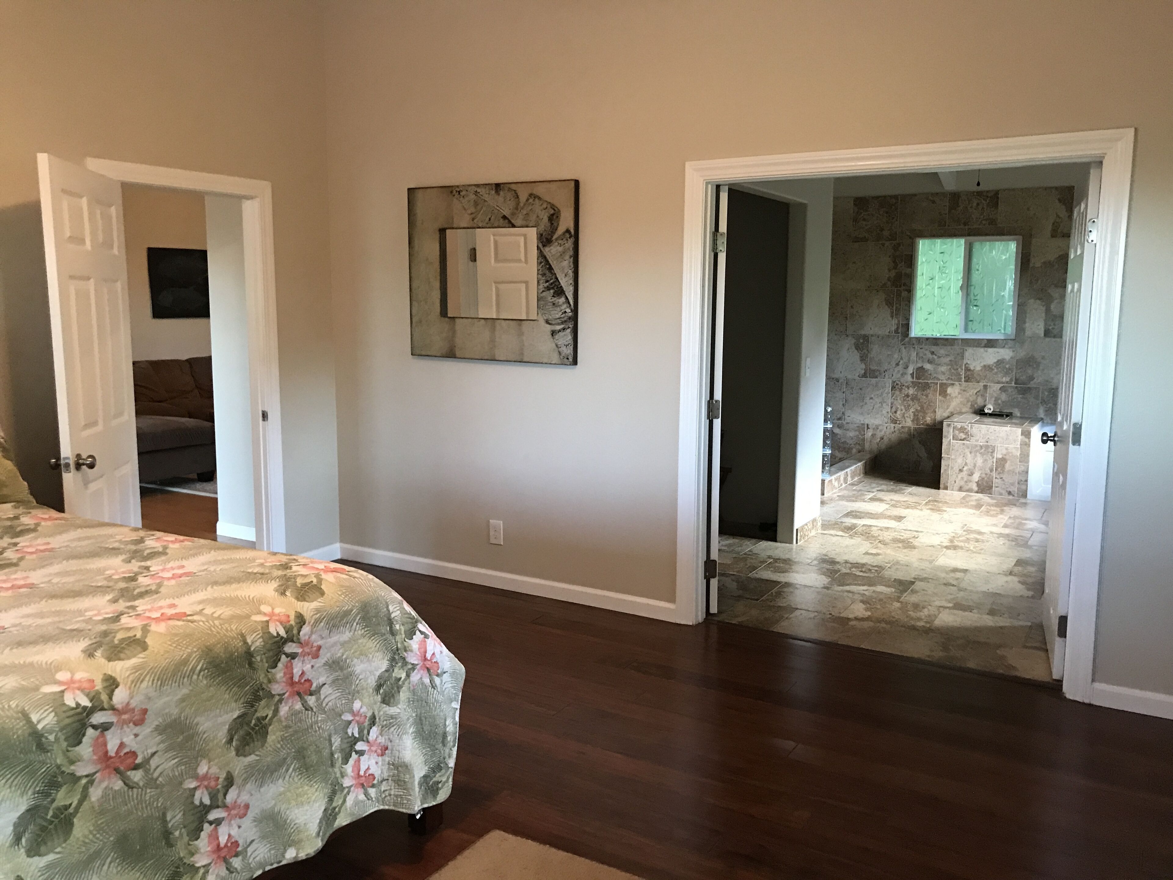 A bedroom with a floral bedspread, a painting on the wall, and an open door leading to a bathroom with a stone wall and a bathtub.