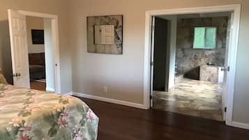 A bedroom with a floral bedspread, a painting on the wall, and an open door leading to a bathroom with a stone wall and a bathtub.
