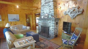 A cozy log cabin interior with a stone fireplace, wooden walls, a sofa, a rocking chair, and a television. - Linekin Vista - 3BDR Waterfront Cottage on Linekin Bay with Private Beach! (East Boothbay)