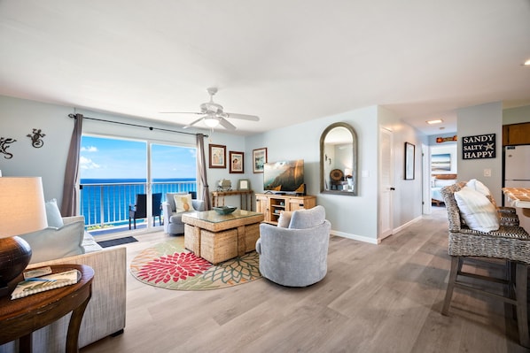 A spacious living room with a large window, a ceiling fan, a television, and a dining area with wicker furniture.