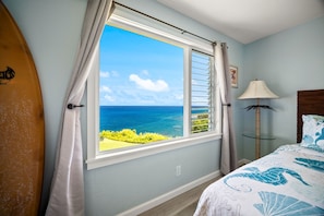 Une chambre dotée d’une grande fenêtre donnant sur l’océan, d’un lit recouvert d’une couvre-lit à motifs floraux bleus et blancs, d’une lampe et d’une planche de surf appuyée contre le mur.