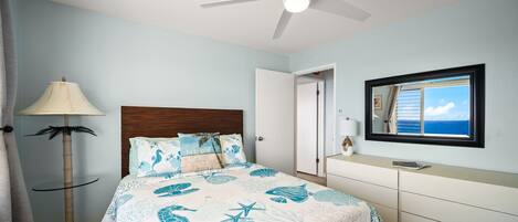 Un dormitorio con cama, ventilador de techo, mesita de noche, cómoda, lámpara y ventana con vistas.