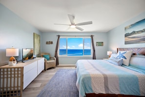 A bedroom with a bed, a desk, a chair, a ceiling fan, a window with a view of the ocean, and a painting on the wall.