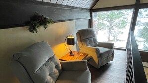 A modern living room with a grey sofa, a small wooden table, and a lamp. There is a window with a view of trees. - River Cottage (South Fork)