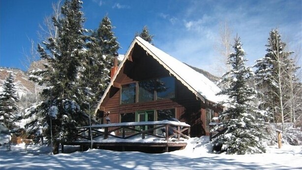 A wooden cabin with a sloped roof, surrounded by snow-covered trees and a clear blue sky. - River Cottage (South Fork)