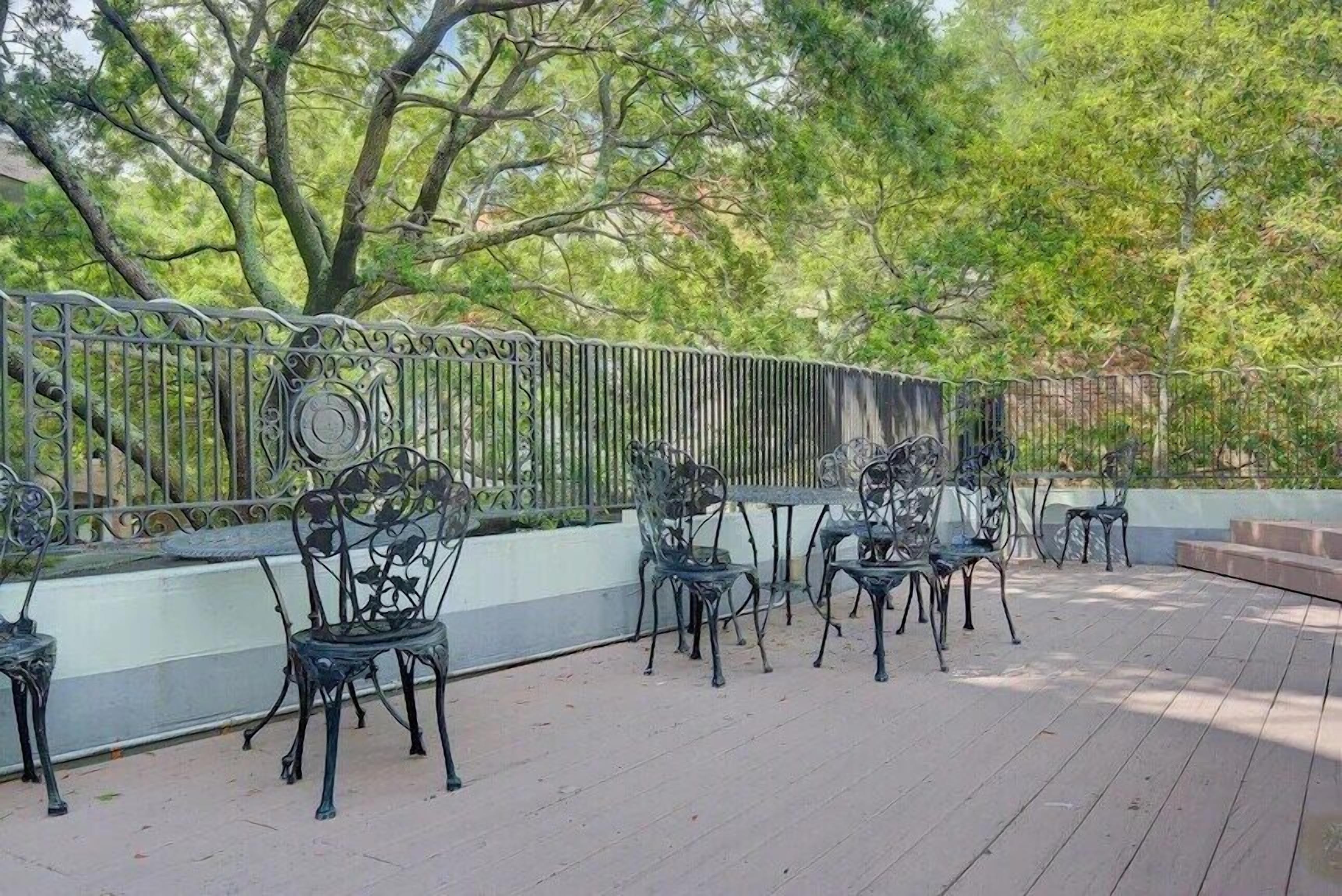 An outdoor patio with metal chairs and tables, surrounded by a metal fence and trees.