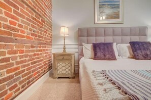A bedroom with a brick wall, a bed with a grey headboard, and a nightstand with a lamp.