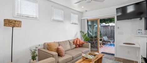 Una sala moderna con un sofá color beige, una mesa de centro de madera y un ventilador de techo. Hay un ventanal corredizo que da a un área exterior con una silla de patio.