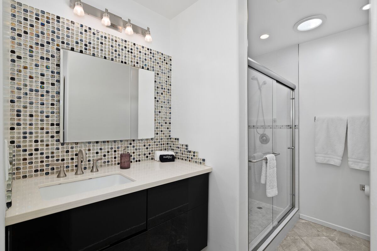 A modern bathroom with a mosaic tile backsplash, a large mirror, a glass shower enclosure, and a white sink with black cabinets.